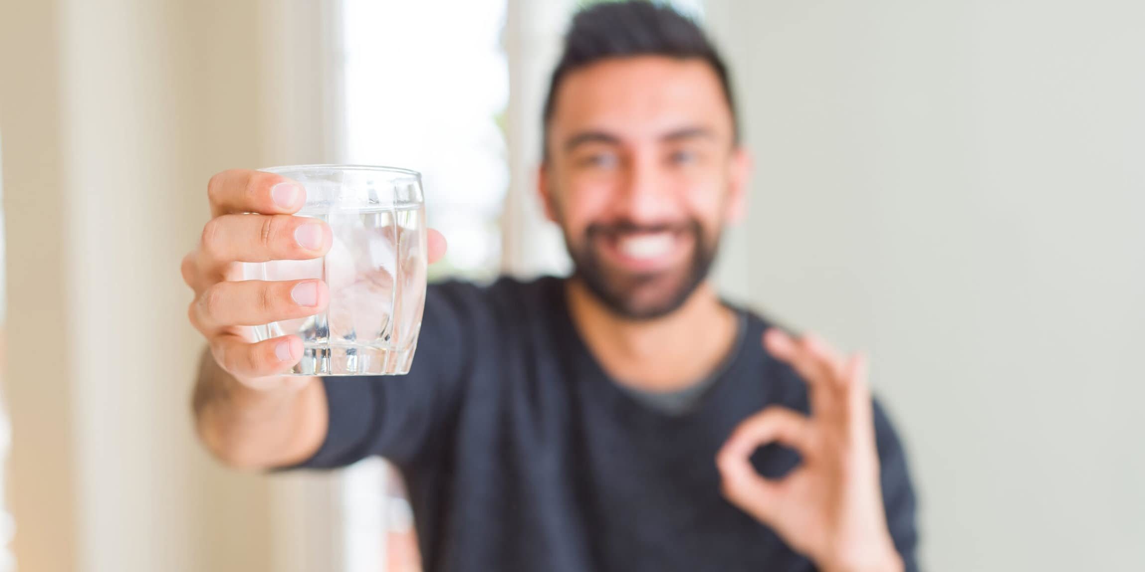 Hübscher hispanischer Mann, der ein frisches Glas Eauvation Wasser trinkt und mit den Fingern ein „OK“-Zeichen macht, ein ausgezeichnetes Symbol